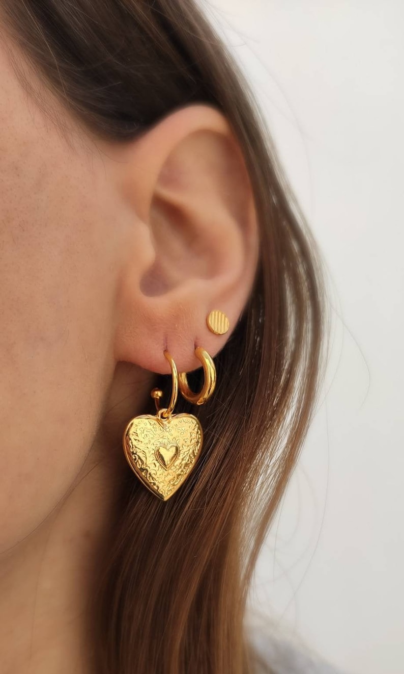 Puede incluir: Un conjunto de pendientes dorados. Un pendiente presenta un colgante en forma de coraz&oacute;n con un dise&ntilde;o en relieve y un coraz&oacute;n m&aacute;s peque&ntilde;o en el centro. Los otros pendientes son un aro peque&ntilde;o y un peque&ntilde;o perno redondo. Los pendientes se llevan en una oreja.