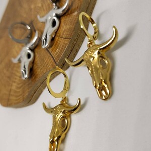 May include: Close-up of gold and silver-colored bull skull earrings. The earrings are detailed with horns and skull features. The gold earrings have a shiny finish, while the silver earrings have a matte finish. The earrings are on a wooden surface.