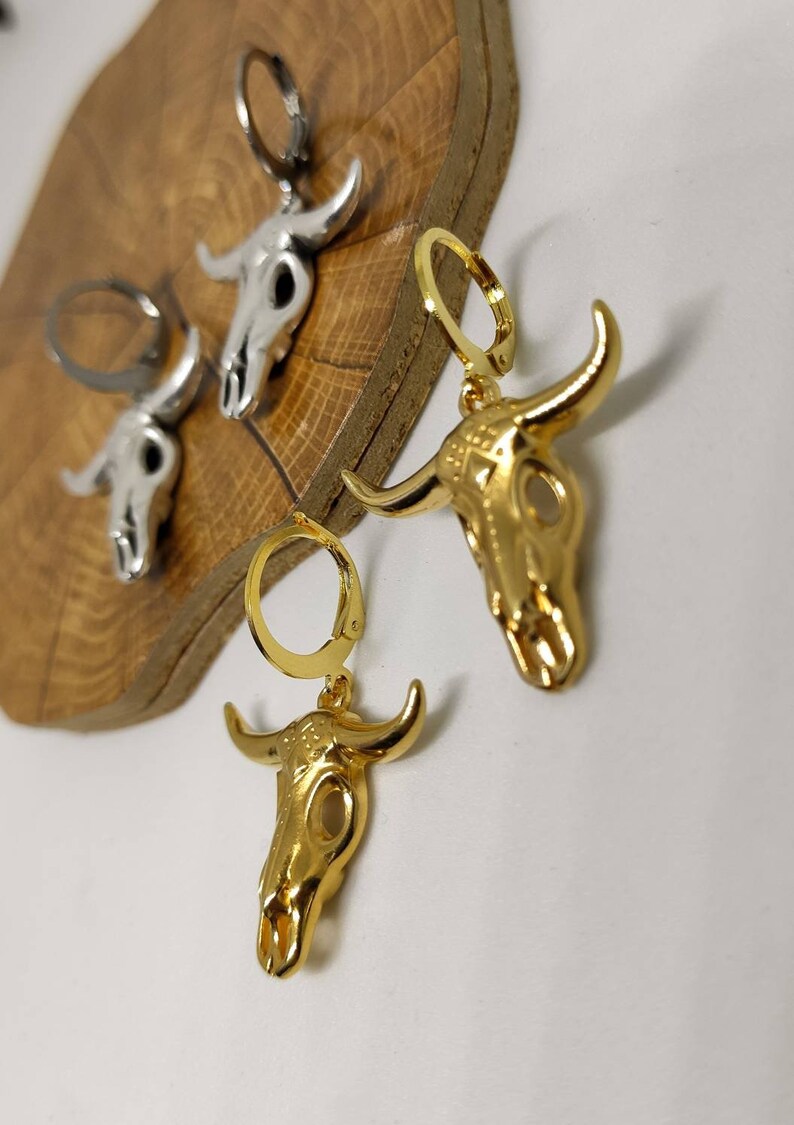 May include: Close-up of gold and silver-colored bull skull earrings. The earrings are detailed with horns and skull features. The gold earrings have a shiny finish, while the silver earrings have a matte finish. The earrings are on a wooden surface.
