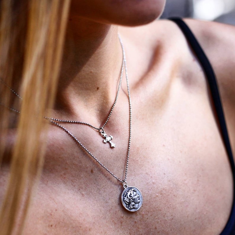 Puede incluir: Un collar plateado de dos capas con dos colgantes. El collar superior tiene un peque&ntilde;o colgante de cruz, y el collar inferior tiene un colgante redondo con un dise&ntilde;o de perfil cl&aacute;sico. Los collares son usados por una persona.