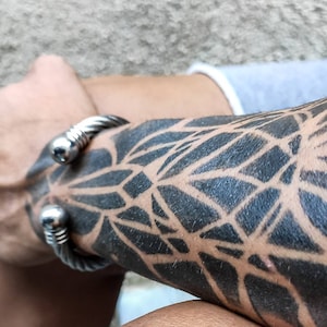 K&ouml;nnte beinhalten: Ein silberfarbenes, gedrehtes Kabelarmband mit abgerundetem, poliertem Ende. Das Armband wird an einem Arm mit einem schwarzen geometrischen Tattoo-Design getragen. Der Arm ist angewinkelt und der Hintergrund ist unscharf.