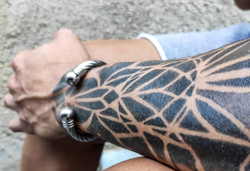 K&ouml;nnte beinhalten: Ein silberfarbenes, gedrehtes Kabelarmband mit abgerundetem, poliertem Ende. Das Armband wird an einem Arm mit einem schwarzen geometrischen Tattoo-Design getragen. Der Arm ist angewinkelt und der Hintergrund ist unscharf.