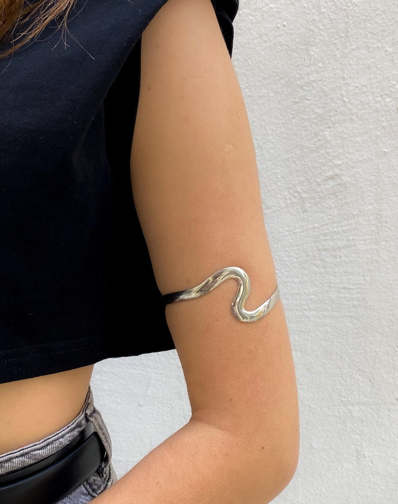 May include: A silver arm cuff with a wavy design. The cuff is worn on the upper arm and has a smooth, polished finish. The jewelry is a simple yet elegant accessory.