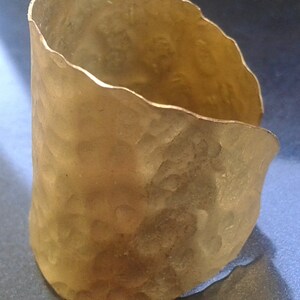 May include: A hammered gold-tone cuff bracelet. The cuff has a textured surface and an irregular, wavy edge. The open cuff design allows for easy wear. The bracelet is a statement piece, suitable for everyday wear or special occasions.