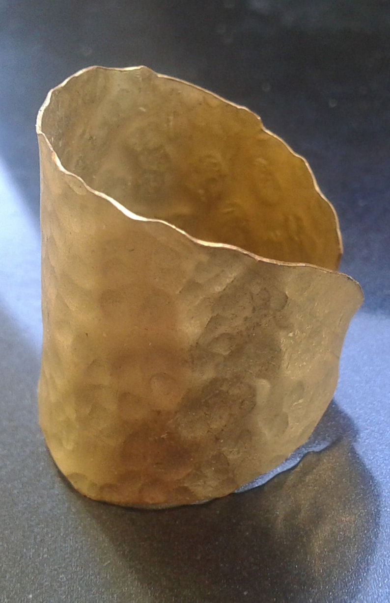 May include: A hammered gold-tone cuff bracelet. The cuff has a textured surface and an irregular, wavy edge. The open cuff design allows for easy wear. The bracelet is a statement piece, suitable for everyday wear or special occasions.