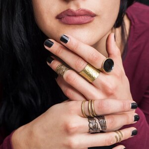 May include: A collection of gold-toned rings of various designs adorn the fingers. The rings include a hammered band, a ring with a black stone, and rings with leaf and spiral patterns. The nails are painted black.