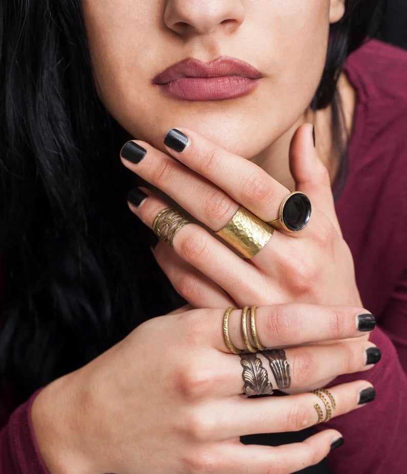 May include: A collection of gold-toned rings of various designs adorn the fingers. The rings include a hammered band, a ring with a black stone, and rings with leaf and spiral patterns. The nails are painted black.