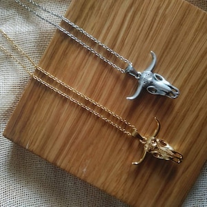 May include: Two necklaces with bull skull pendants. One necklace is gold-colored, the other is silver-colored. Both pendants have detailed skull features and are on delicate chains. The necklaces rest on a wooden surface.