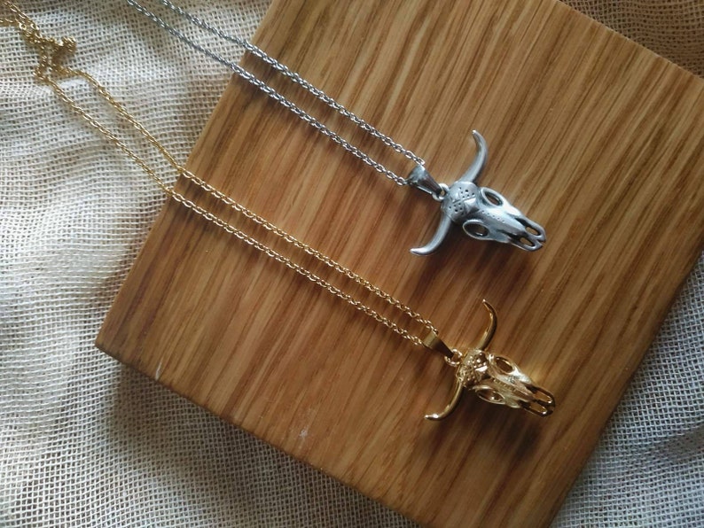 May include: Two necklaces with bull skull pendants. One necklace is gold-colored, the other is silver-colored. Both pendants have detailed skull features and are on delicate chains. The necklaces rest on a wooden surface.