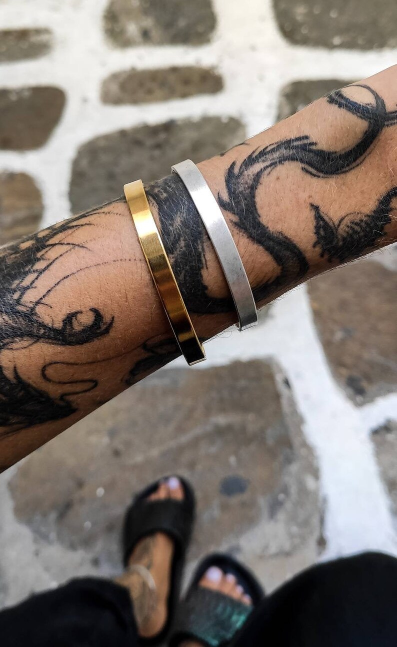 Pode incluir: S&atilde;o exibidas duas pulseiras minimalistas, uma dourada e outra prateada. A pulseira dourada est&aacute; &agrave; esquerda e a prateada &agrave; direita. As pulseiras s&atilde;o usadas em um bra&ccedil;o com uma grande tatuagem preta. O fundo &eacute; uma superf&iacute;cie de pedra branca e cinza.