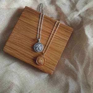 Puede incluir: Dos collares con colgantes de monedas sobre una superficie de madera. Uno es plateado con un perfil en la moneda, el otro dorado con un dise&ntilde;o de sol. El fondo es de tela clara. Los collares son de metal.