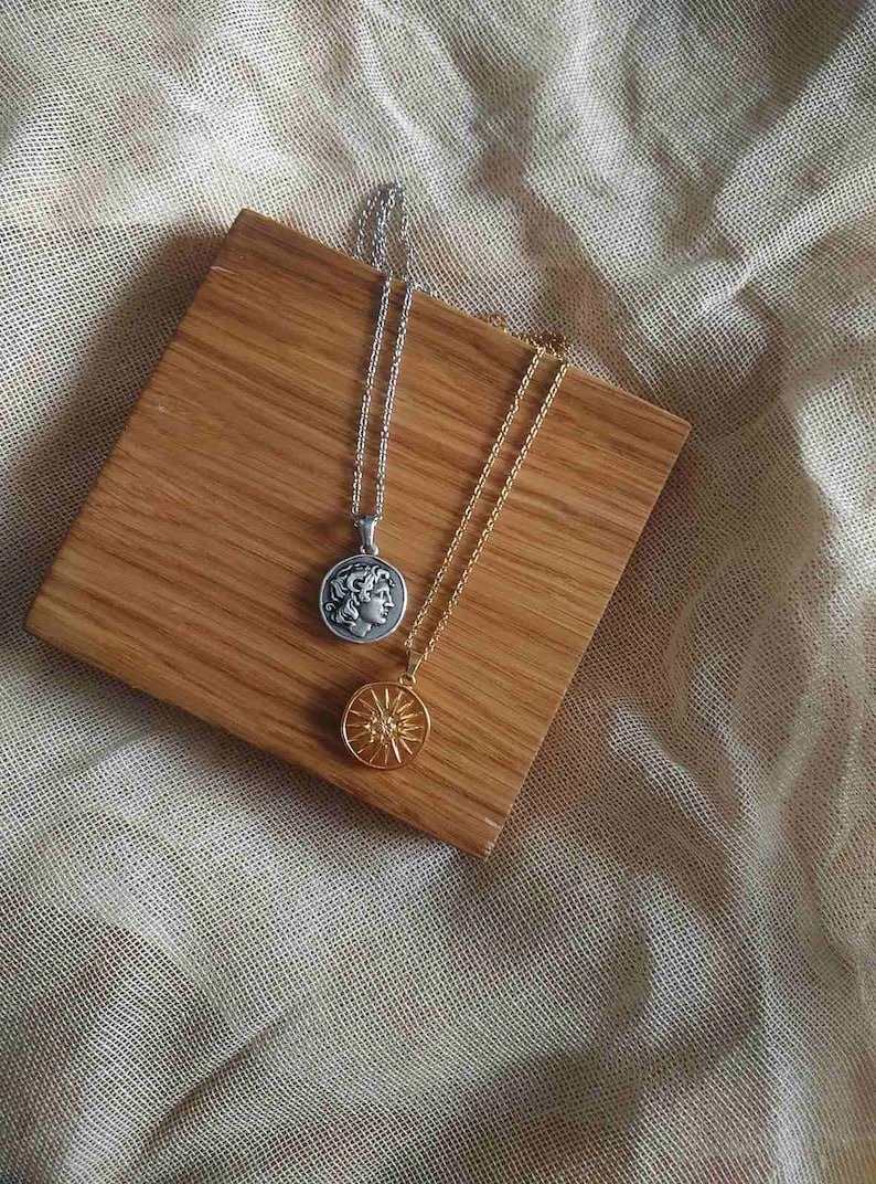 Puede incluir: Dos collares con colgantes de monedas sobre una superficie de madera. Uno es plateado con un perfil en la moneda, el otro dorado con un dise&ntilde;o de sol. El fondo es de tela clara. Los collares son de metal.
