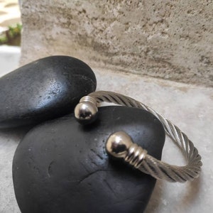 K&ouml;nnte beinhalten: Ein silberfarbenes Armband aus gedrehtem Kabel mit abgerundeten Endkappen. Das Armband wird auf einem schwarzen, glatten Stein pr&auml;sentiert, mit einem weiteren Stein im Hintergrund. Das Armband hat ein strukturiertes, geflochtenes Aussehen.