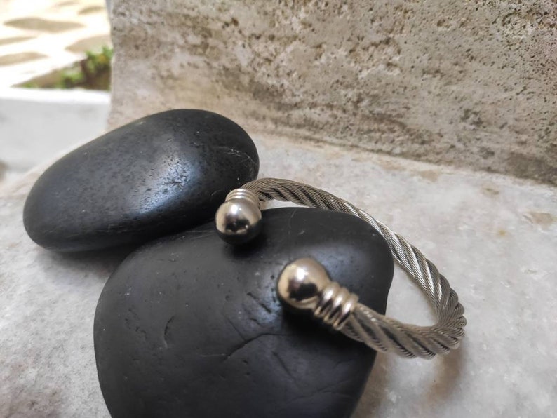 K&ouml;nnte beinhalten: Ein silberfarbenes Armband aus gedrehtem Kabel mit abgerundeten Endkappen. Das Armband wird auf einem schwarzen, glatten Stein pr&auml;sentiert, mit einem weiteren Stein im Hintergrund. Das Armband hat ein strukturiertes, geflochtenes Aussehen.