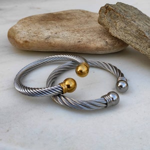 K&ouml;nnte beinhalten: Zwei silberfarbene, gedrehte Kabelarmb&auml;nder mit goldenen und silbernen Kugelakzenten. Die Armb&auml;nder sind offen. Der Schmuck wird auf einer wei&szlig;en Oberfl&auml;che mit einem Naturstein im Hintergrund pr&auml;sentiert.