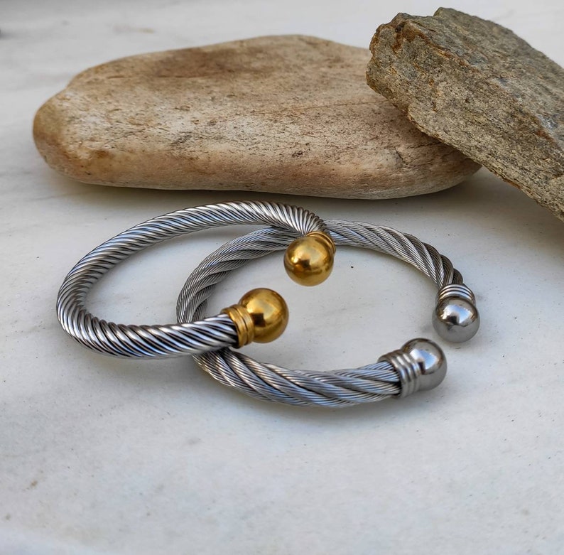 K&ouml;nnte beinhalten: Zwei silberfarbene, gedrehte Kabelarmb&auml;nder mit goldenen und silbernen Kugelakzenten. Die Armb&auml;nder sind offen. Der Schmuck wird auf einer wei&szlig;en Oberfl&auml;che mit einem Naturstein im Hintergrund pr&auml;sentiert.