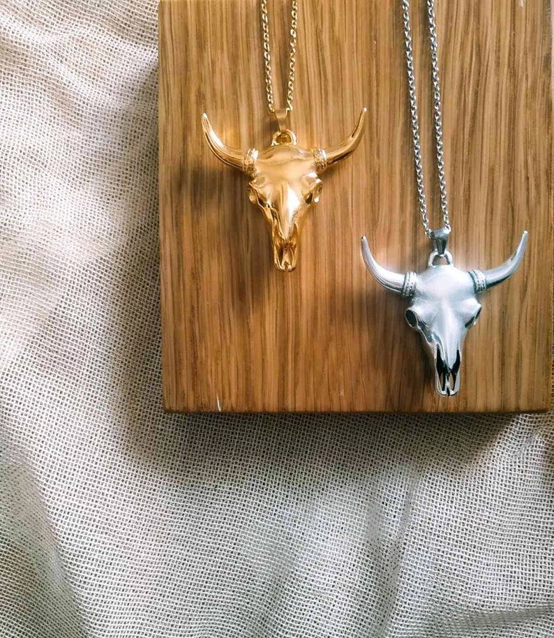 May include: Two silver chains with bull skull pendants. The pendant on the left chain is gold, and the pendant on the right chain is silver. The chains are displayed on a wooden surface.