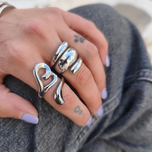 May include: Two polished silver-toned rings worn on the fingers. One ring features an open, fluid heart-shaped design, while the other ring has a thick, smooth, multi-layered band with a rounded, organic shape that wraps around the finger.