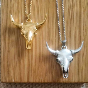 May include: Two silver and gold bull skull pendants on chains. The pendants are on a wooden background.