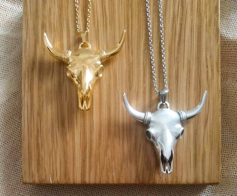 May include: Two silver and gold bull skull pendants on chains. The pendants are on a wooden background.
