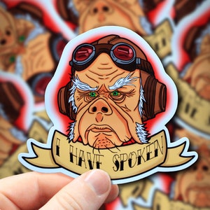May include: A sticker featuring a cartoon character with a large nose, goggles, and a white beard. The character is wearing a brown pilot's helmet and has green eyes. The sticker has a yellow banner with the text "I Have Spoken" in a vintage style font.