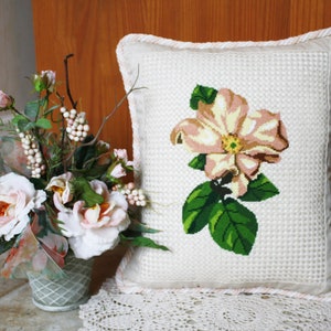 May include: A white pillow with a pink flower embroidered on it. The pillow has a white and pink trim around the edges. The pillow is sitting on a white lace doily.
