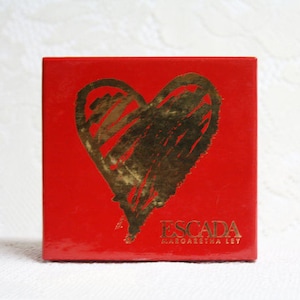 May include: A red box with a gold heart design. The box has the brand name "ESCADA" and "MARGARETHA LEY" printed in gold. The heart is a rough, textured design.