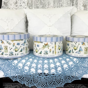 May include: Three round decorative boxes with lids. The boxes are white with a blue striped border and feature illustrations of butterflies, watering cans, and birds. The boxes are arranged on a blue lace doily.