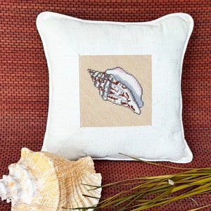 May include: A white pillow with a beige square in the center featuring an embroidered seashell. The seashell is brown, white, and pink. The pillow is on a brown woven surface.