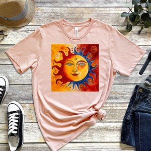 May include: A pink t-shirt featuring a colourful sun graphic. The sun has a face with blue eyes and a yellow smile. The background of the sun is a mix of orange, red and yellow.