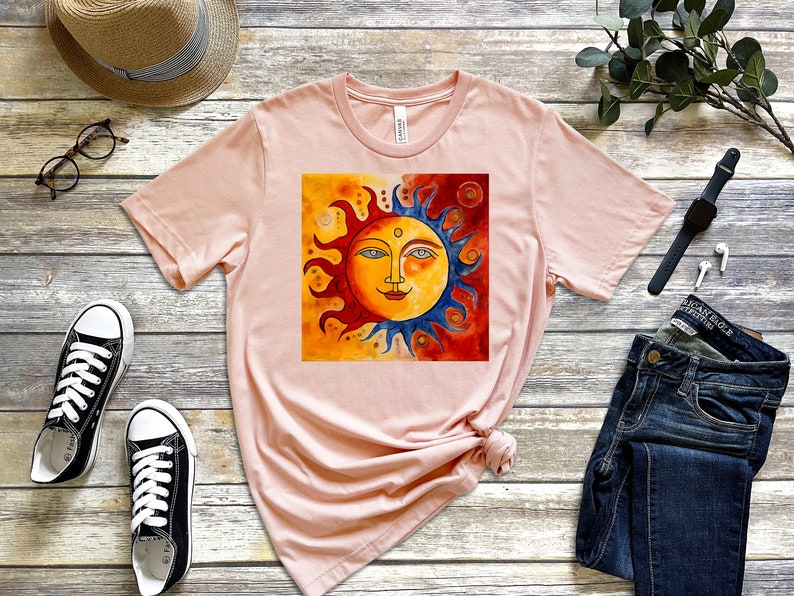 May include: A pink t-shirt featuring a colourful sun graphic. The sun has a face with blue eyes and a yellow smile. The background of the sun is a mix of orange, red and yellow.