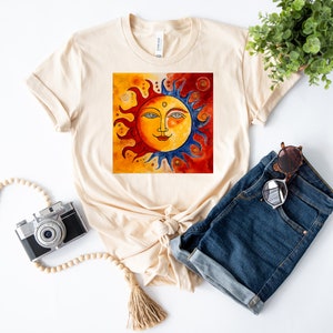 May include: A beige t-shirt featuring a graphic of a stylised sun with a face, surrounded by red, orange and blue flames. The sun is set against a background of orange, red and yellow.