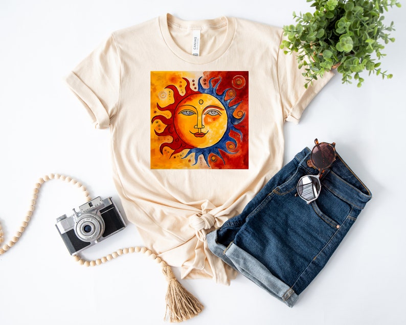 May include: A beige t-shirt featuring a graphic of a stylised sun with a face, surrounded by red, orange and blue flames. The sun is set against a background of orange, red and yellow.