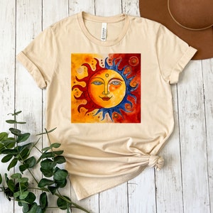 May include: A beige t-shirt featuring a colourful sun and moon design. The sun has a smiling face and is surrounded by yellow, orange and red rays. The moon is blue and has a white crescent shape.