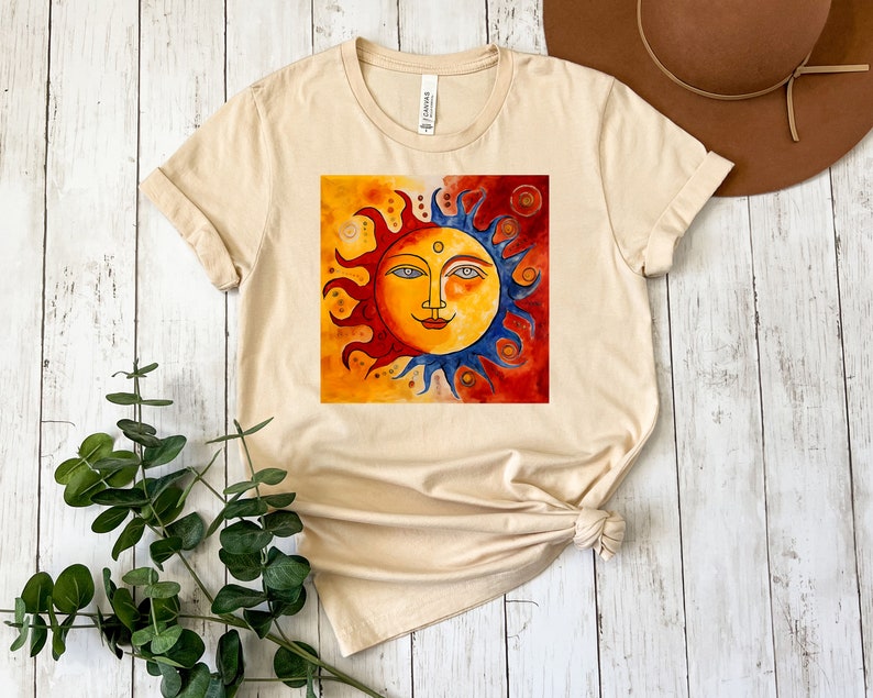 May include: A beige t-shirt featuring a colourful sun and moon design. The sun has a smiling face and is surrounded by yellow, orange and red rays. The moon is blue and has a white crescent shape.