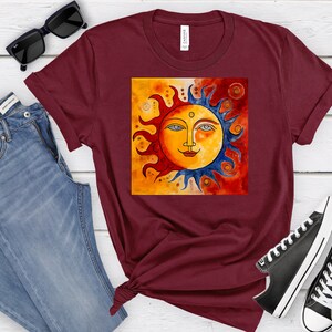 May include: A maroon t-shirt featuring a graphic of a stylised sun with a face. The sun is painted in shades of yellow, orange, red, and blue. The background is a gradient of orange and red.