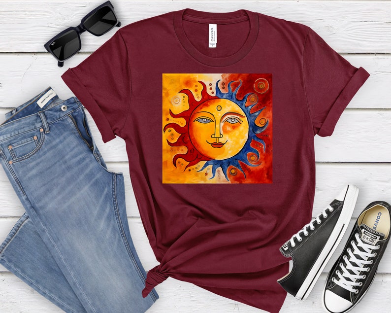 May include: A maroon t-shirt featuring a graphic of a stylised sun with a face. The sun is painted in shades of yellow, orange, red, and blue. The background is a gradient of orange and red.