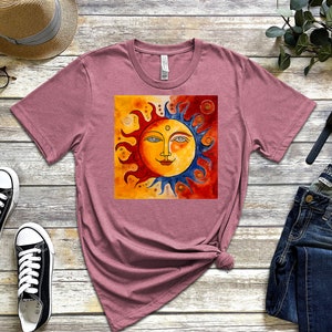 May include: A mauve t-shirt featuring a graphic of a stylised sun with a smiling face. The sun is half yellow and half blue, with red and orange flames. The background is a watercolour wash of yellow, orange, and red.
