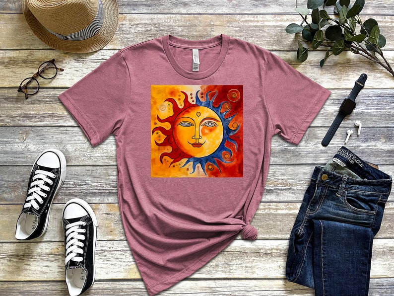 May include: A mauve t-shirt featuring a graphic of a stylised sun with a smiling face. The sun is half yellow and half blue, with red and orange flames. The background is a watercolour wash of yellow, orange, and red.