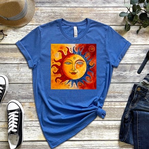 May include: A blue t-shirt featuring a graphic of a stylised sun with a face. The sun is half orange and half blue, with a red background. The t-shirt is laid flat on a wooden surface.