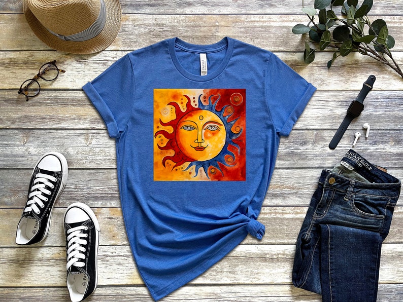 May include: A blue t-shirt featuring a graphic of a stylised sun with a face. The sun is half orange and half blue, with a red background. The t-shirt is laid flat on a wooden surface.