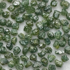 Op de afbeelding: Een verzameling ruwe, ongeslepen groene diamanten. De edelstenen variëren in grootte en vorm en vertonen een reeks groene tinten met enkele hints van blauw en zilver. De diamanten zijn verspreid over een wit oppervlak.