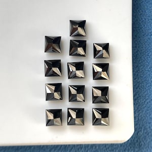 Könnte beinhalten: Eine Ansammlung von dreizehn kleinen, quadratischen schwarzen Edelsteinen, die in einem Raster angeordnet sind. Jeder Stein weist eine facettierte, pyramidenartige Oberseite mit scharfen Kanten und reflektierenden Flächen auf, die ein geometrisches Muster bilden.