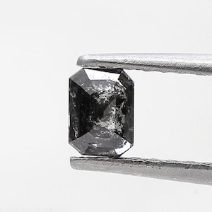 Puede incluir: Un diamante negro de talla rectangular, sujeto con unas pinzas plateadas. La gema tiene un aspecto moteado y una superficie pulida. El diamante se encuentra sobre un fondo blanco liso. Medidas en milímetros.