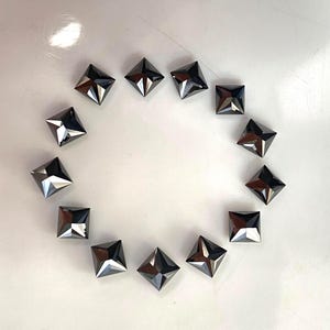 May include: A collection of twelve dark gray, square-shaped faceted crystals arranged in a circular pattern. Each crystal features a pyramid-like top with sharp, clean edges and a reflective, metallic-like surface that catches the light.