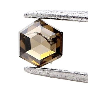 May include: A hexagonal-cut, brown diamond held by silver tweezers. The gemstone has a faceted surface with a visible crack. The diamond's color transitions from light brown to a golden hue.