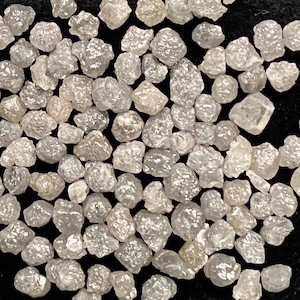 May include: A collection of small, rough-cut diamonds displayed in a black velvet-lined metal case. The diamonds are various sizes and shapes, with a light gray color and a slightly frosted appearance.