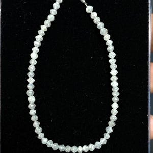 May include: A delicate bracelet composed of numerous small, faceted, light gray gemstones. The stones are strung together on a thin, silver-colored chain, creating a sparkling, elegant accessory. The bracelet is displayed against a dark background.