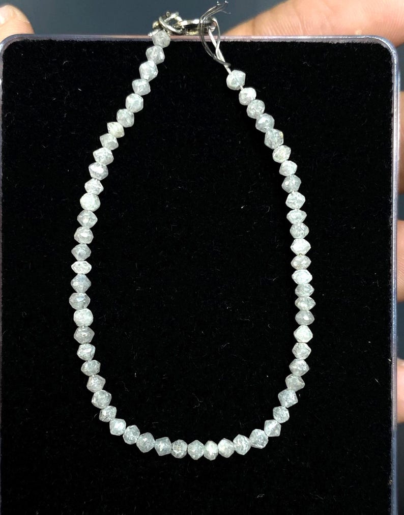 May include: A delicate bracelet composed of numerous small, faceted, light gray gemstones. The stones are strung together on a thin, silver-colored chain, creating a sparkling, elegant accessory. The bracelet is displayed against a dark background.
