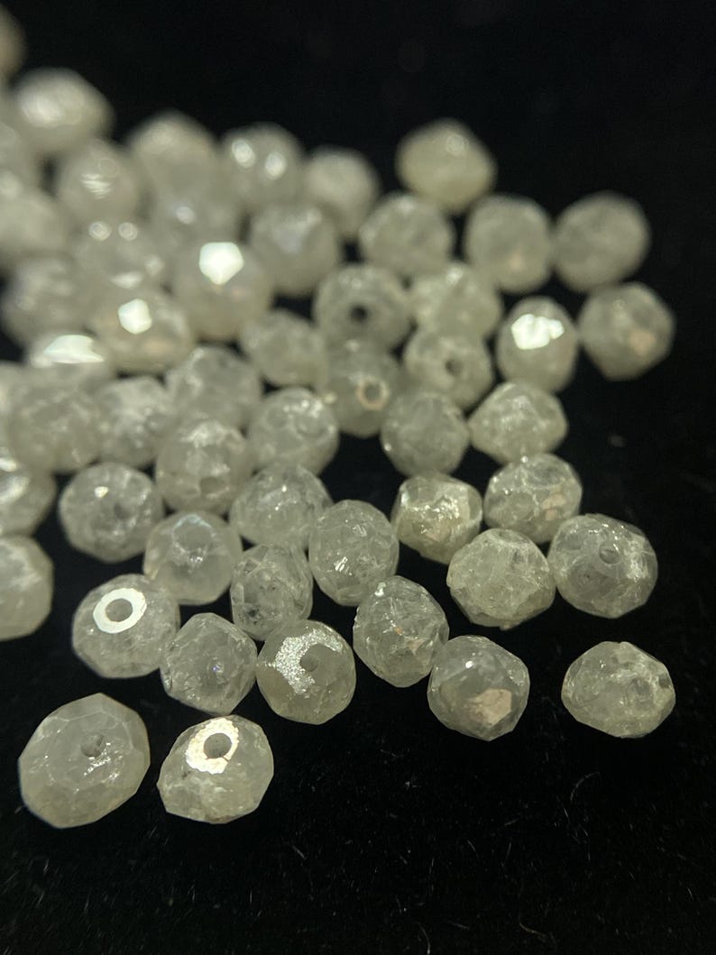 May include: A collection of small, faceted, clear beads with a slightly iridescent sheen. The beads are irregularly shaped and appear to be made of a translucent material, possibly glass or crystal. They are scattered against a dark background.